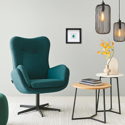 Quadratischer schwarzer Bilderrahmen mit einem weißen Passepartout rahmt einen türkisen Vulva-Abdruck, der mit Gouachefarbe auf Aquarellkarton gefertigt wurde. Das Bild hängt über einem türkisen Sessel. Rechts von dem Sessel sind zwei Beistelltische mit Büchern und einer Vase dekoriert. Über den Beistelltischen hängen zwei Drahtgeflecht-Lampen.