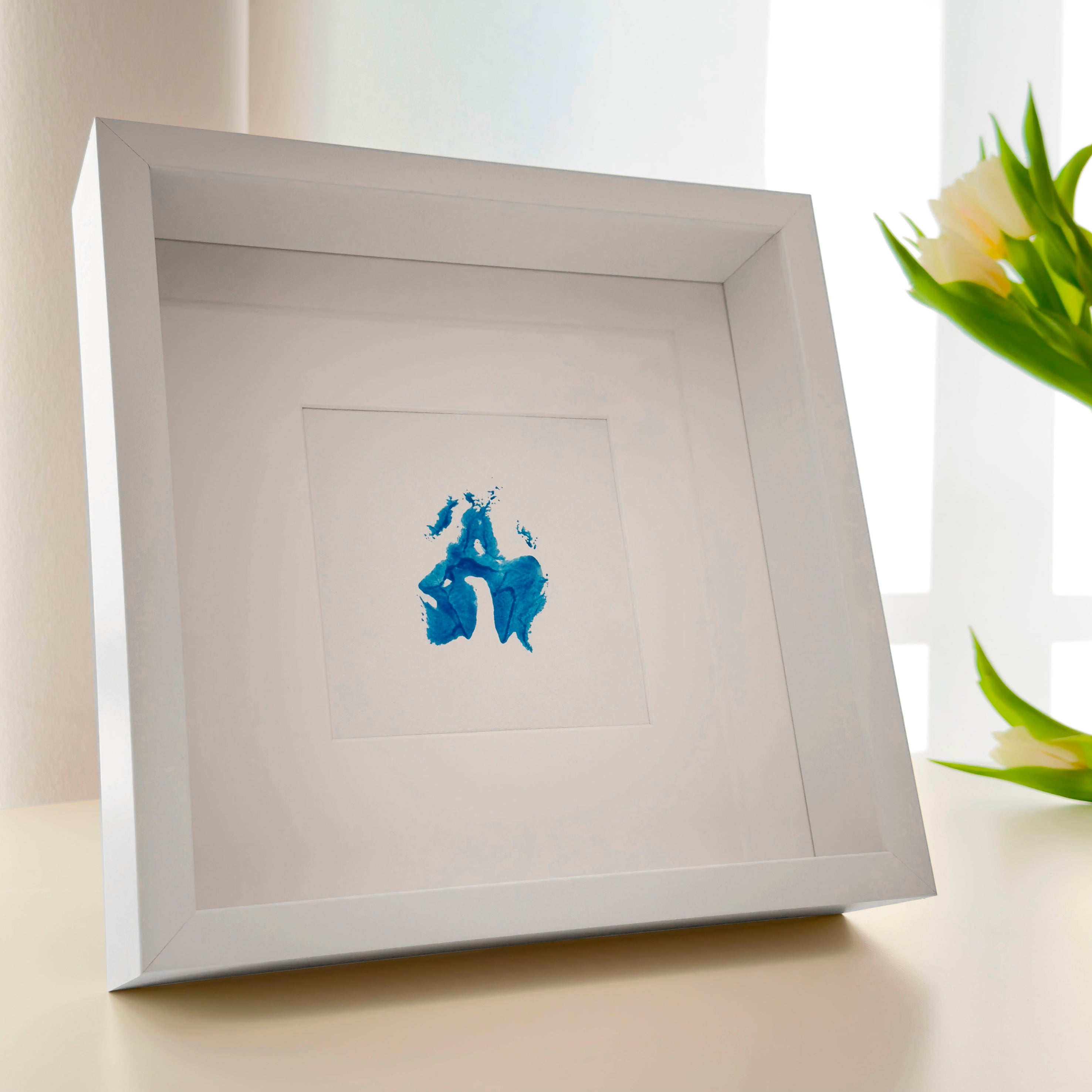 Perspektivische Aufnahme von quadratischem weißer Bilderrahmen mit Passepartout und blauer Vulva, die mit Gouachefarbe auf Aquarellkarton gestempelt wurde. Hintergrund Fenster und Tulpen
