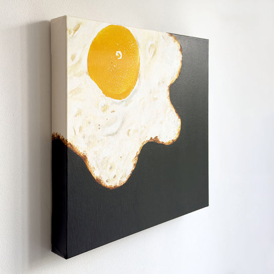 Perspektivische Aufnahmen vom Kunstwerk BC #06 Sonny Side Up. Das Kunstwerk ist 40x40cm und zeigt ein Spiegelei, welches von oben links ins Bild fließt. Das Eigelb wurde mit dem Abdruck der Brust der Künstlerin gefertigt. Der Hintergrund ist schwarz. Das Spiegelei fließt auch über den linken und oberen Rand.